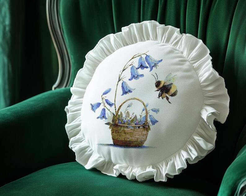 May include: A round white pillow with a ruffled edge, featuring a floral design of bluebell flowers and a bee. The flowers are in a woven basket. The pillow is set against a green velvet chair.