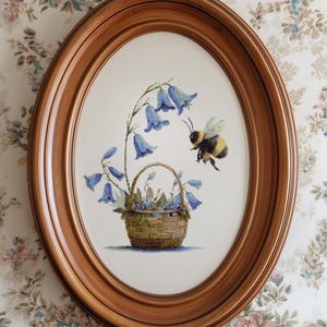 May include: Oval wooden framed cross-stitch art featuring a basket of bluebell flowers and a bumblebee. The artwork is set against a white background and framed in a warm, medium-toned wood. The floral design is detailed and colorful.