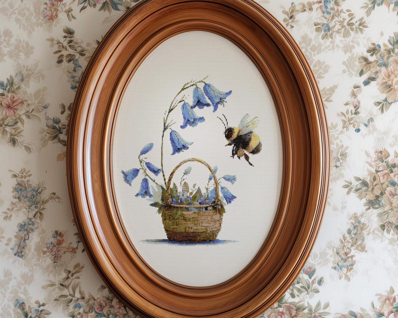 May include: Oval wooden framed cross-stitch art featuring a basket of bluebell flowers and a bumblebee. The artwork is set against a white background and framed in a warm, medium-toned wood. The floral design is detailed and colorful.