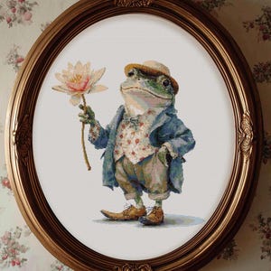 May include: Cross-stitch artwork of a frog dressed in a blue jacket, vest, and hat, holding a flower. The frog is in a gold oval frame. The background is a floral wallpaper.