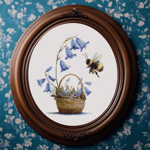 May include: A framed cross-stitch artwork featuring a basket of bluebell flowers and a bumblebee. The artwork is set in a round, dark brown wooden frame. The background is a white canvas. The bee is in flight, approaching the flowers.