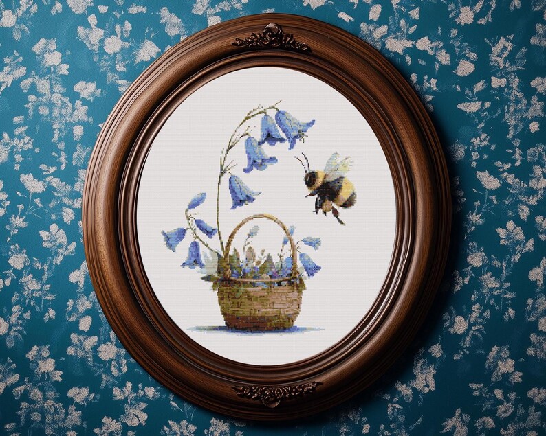 May include: A framed cross-stitch artwork featuring a basket of bluebell flowers and a bumblebee. The artwork is set in a round, dark brown wooden frame. The background is a white canvas. The bee is in flight, approaching the flowers.