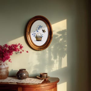 May include: An oval, gold-framed artwork depicts a basket of bluebell flowers with a bee. The artwork hangs on a light green wall. A wooden table displays a vase of pink flowers, a small dark vase, and a decorative plate.