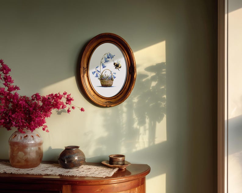 May include: An oval, gold-framed artwork depicts a basket of bluebell flowers with a bee. The artwork hangs on a light green wall. A wooden table displays a vase of pink flowers, a small dark vase, and a decorative plate.