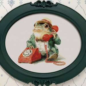 Könnte beinhalten: Ein gerahmtes Kunstwerk mit einem Frosch in Anzug und Hut, der einen roten Telefonhörer ans Ohr hält. Der Frosch sitzt neben einem roten Wählscheibentelefon. Der Rahmen ist dunkelgrün und oval.