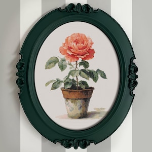 以下が含まれることがあります： 鉢植えのバラの楕円形の額入りプリント。バラは鮮やかなコーラル色で、緑の葉と茶色の鉢があります。フレームは濃い緑色で、装飾的なディテールがあります。背景は白とグレーのストライプ模様です。