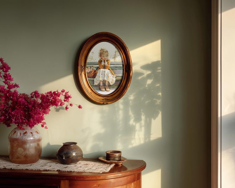 May include: An oval framed print of a young girl in a dress, seated with a basket. The frame is a warm brown color, and the print is mounted on a light green wall. A bouquet of pink flowers in a ceramic vase sits on a wooden table.