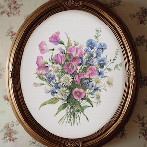 May include: An oval, gold-framed cross-stitch of a floral bouquet. The bouquet features pink, purple, blue, and white flowers with green foliage. The frame has ornate detailing. The background is a cream-colored fabric.