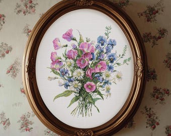 May include: An oval, gold-framed cross-stitch of a floral bouquet. The bouquet features pink, purple, blue, and white flowers with green foliage. The frame has ornate detailing. The background is a cream-colored fabric.