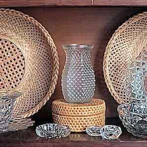 Op de afbeelding: Een plank met een verscheidenheid aan glazen en rieten voorwerpen. Er zijn twee rieten manden, twee glazen vazen, twee glazen flessen, een glazen kom en een glazen pot. De voorwerpen zijn decoratief gerangschikt.