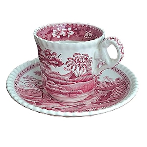 May include: A white teacup and saucer set with a red scenic design. The cup and saucer feature a detailed landscape with trees, mountains, and a volcano. The cup has a decorative handle and a scalloped rim. The saucer has a matching design and a ridged edge.