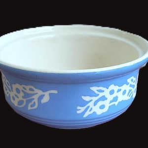 May include: A round ceramic bowl featuring a light blue exterior with a white floral vine pattern wrapping around the sides. The interior of the bowl is a solid off-white color, and the rim has a subtle, textured, ridged edge detail.