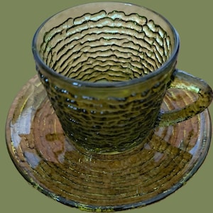 May include: A green glass teacup and saucer set with a textured, bumpy surface. The cup has a handle and the saucer has a slightly raised edge.