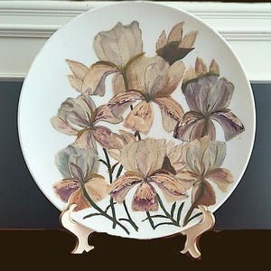 May include: A white ceramic plate featuring a hand-painted floral design of irises in shades of beige, purple, and brown. The plate is displayed on a gold-colored stand, showcasing the artwork. The flowers have green stems and leaves.
