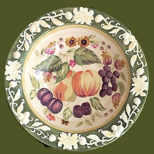 May include: A round, decorative ceramic bowl with a hand-painted fruit design. The bowl features peaches, plums, grapes, and flowers in shades of orange, purple, and pink. The rim is green with white floral accents.