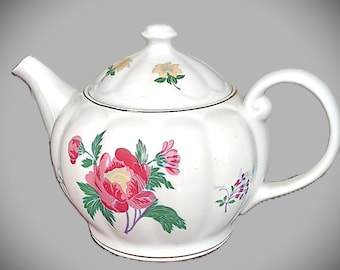 May include: White ceramic teapot with a lid, featuring a large pink peony floral design with green leaves. The teapot has a curved handle and spout, and is trimmed with a gold band. The lid is adorned with small yellow floral accents.