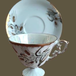 Könnte beinhalten: Ein Vintage-Tee-Set. Die Tasse und Untertasse sind weiß mit goldenen Verzierungen und weisen ein Weizen- und Blumendesign in Braun und Grau auf. Die Tasse hat einen Sockel und einen verzierten Griff.
