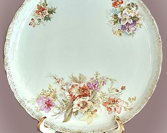 May include: A round, light blue ceramic plate with a scalloped edge and gold trim. The plate features floral arrangements in shades of pink, orange, and purple. The plate is displayed on a gold-colored stand.