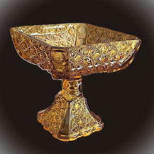May include: A decorative amber-colored glass bowl with a square shape and a pedestal base. The bowl features a detailed, geometric pattern throughout, creating a textured appearance. The base is also intricately designed, adding to the overall elegance of the piece.