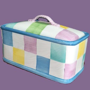 May include: A rectangular ceramic storage box with a pastel checkered pattern in blue, yellow, pink, and green. The lid has a matching pattern and a pink rope handle. The box has a blue trim.