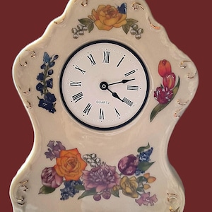 May include: Cream-colored ceramic mantel clock with a floral design. The clock face has black Roman numerals and the word "QUARTZ". The clock is decorated with orange, yellow, purple, and blue flowers.