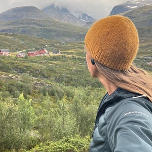 Pode incluir: Uma pessoa usando um gorro de malha amarelo mostarda e uma jaqueta verde escuro, olhando para uma paisagem montanhosa. O fundo apresenta colinas verdes, árvores e edifícios vermelhos sob um céu nublado. A jaqueta tem a palavra "ENDURANCE" impressa.