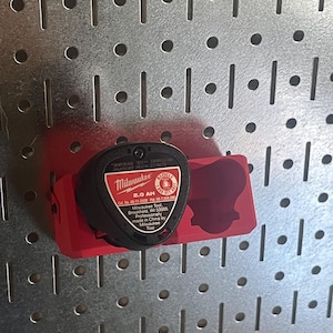 May include: A red plastic battery mount attached to a metal pegboard, holding a black Milwaukee 12V 2.0 AH lithium-ion battery. The battery label displays text including Milwaukee, 2.0 AH, 12V, and manufacturing information from Brookfield, WI.