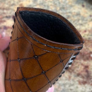 May include: A brown leather card holder with a geometric pattern and black stitching. The interior is black. The edges are decorated with small, dark studs. The card holder is held in a hand.