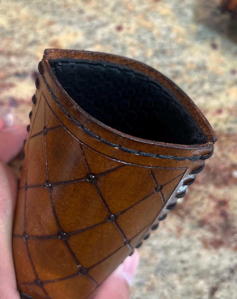 May include: A brown leather card holder with a geometric pattern and black stitching. The interior is black. The edges are decorated with small, dark studs. The card holder is held in a hand.