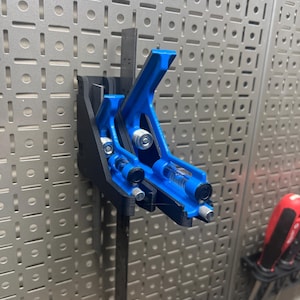 May include: A blue and black combination square tool with a level and ruler attached, mounted on a metal pegboard. The tool is designed for measuring and marking angles and lengths. The background features a silver pegboard with a grid pattern.