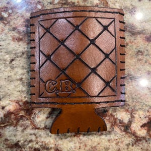 May include: A brown leather beverage holder with a diamond pattern stitched in black thread. The holder has a base and is stitched around the edges. The initials "C.B." are embossed on the front.