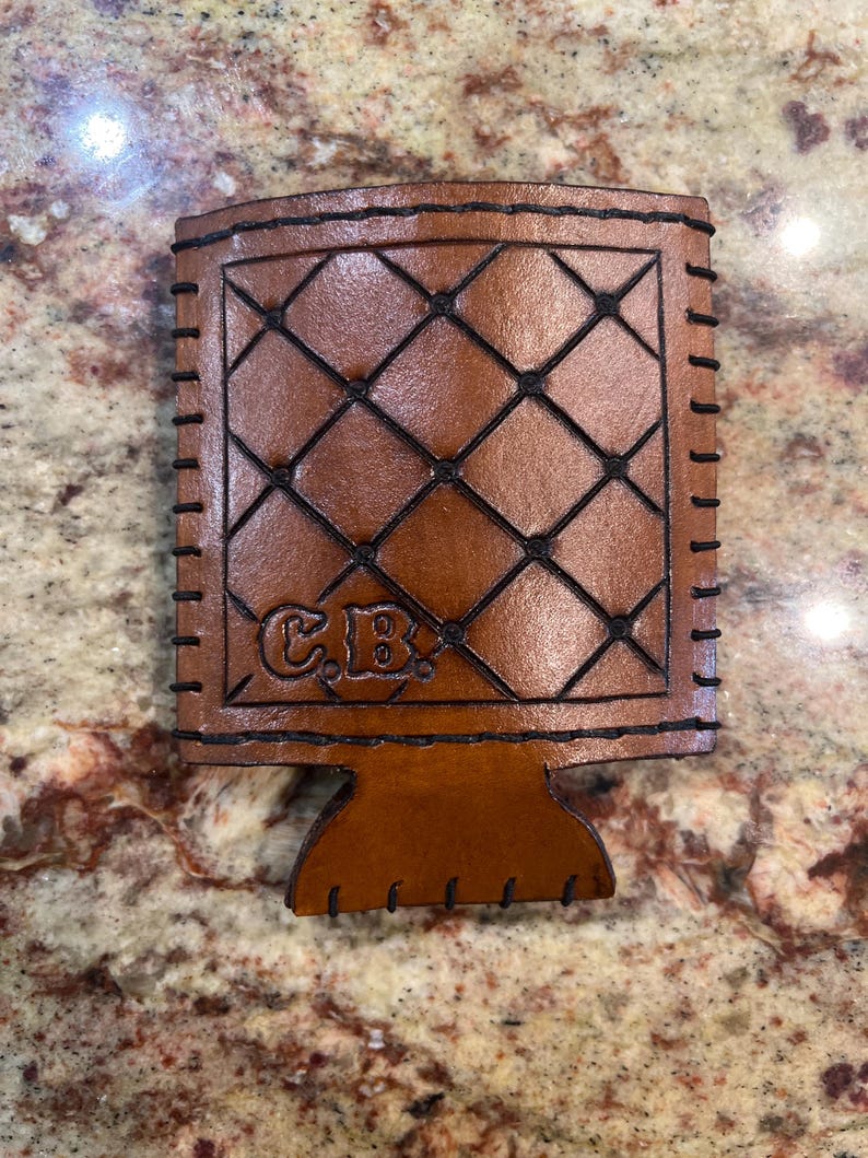 May include: A brown leather beverage holder with a diamond pattern stitched in black thread. The holder has a base and is stitched around the edges. The initials "C.B." are embossed on the front.