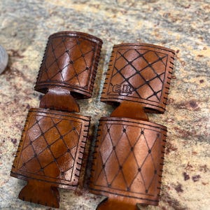 May include: Four brown leather drink holders with a diamond pattern and stitched edges. One holder has the initials "C.B." embossed on it. The holders are designed to keep beverages cold and are made of a durable material.