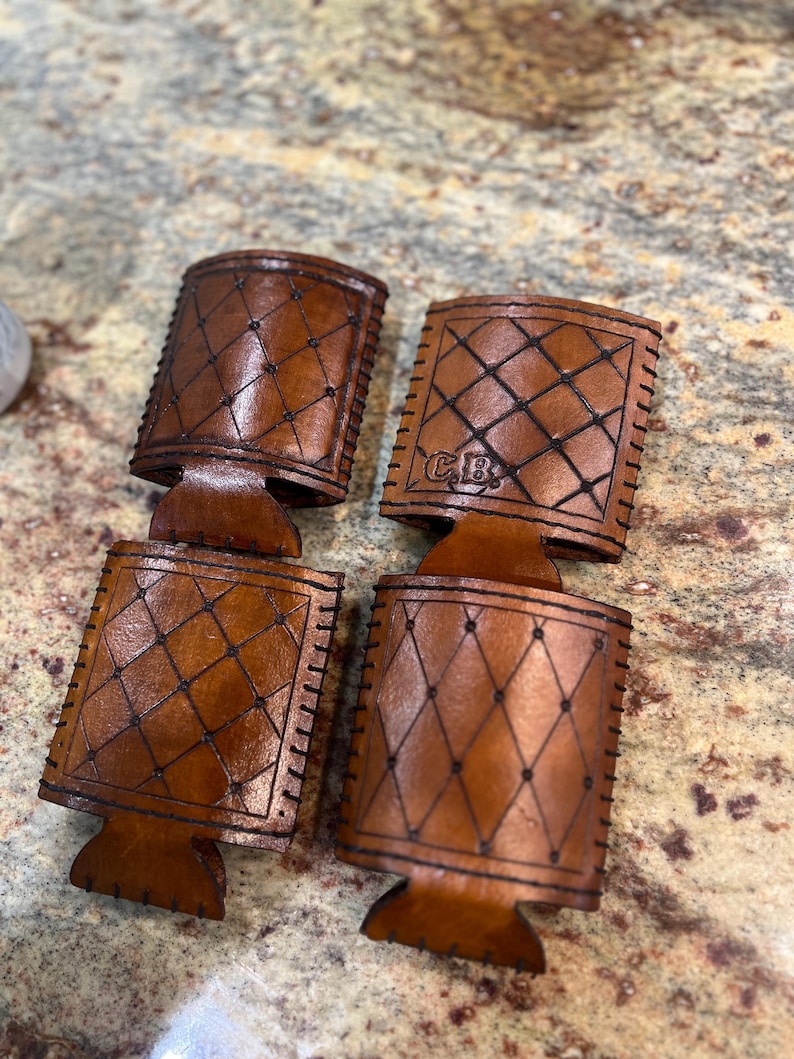May include: Four brown leather drink holders with a diamond pattern and stitched edges. One holder has the initials "C.B." embossed on it. The holders are designed to keep beverages cold and are made of a durable material.
