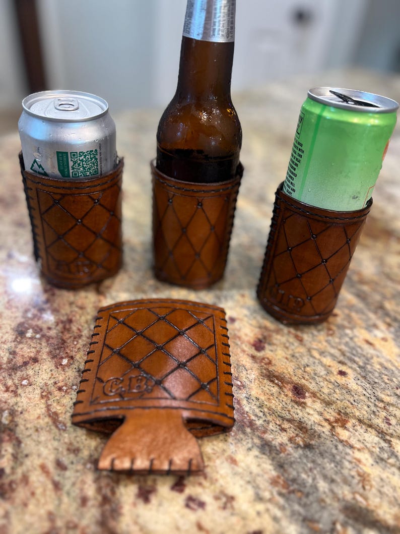 May include: A set of handcrafted brown leather drink holders and a matching coaster. The holders feature a diamond pattern and are designed to hold a beer bottle and cans. The coaster has the initials "C.B." embossed on it.