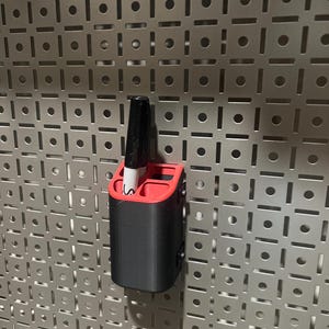 May include: A black and red pen holder with a black marker inside, mounted on a silver pegboard. The pen holder is rectangular with a red top and a black body. The marker is black with a white tip.