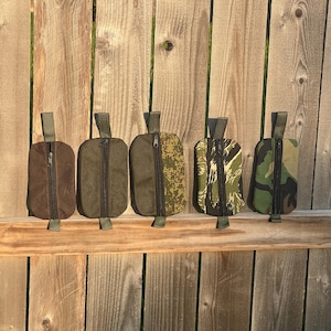 May include: Five small, rectangular pouches with a zipper closure and a fabric loop at the top. The pouches are in various colors and patterns, including solid brown, olive green, and camouflage. They are displayed against a wooden fence.