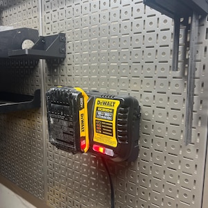 May include: A black and yellow DeWalt DCB1104 12V/20V lithium-ion battery charger with a red charging indicator light. The charger is mounted on a perforated metal wall. A black battery is attached to the charger.
