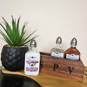 May include: Three glass bottles with silver tops and labels that say "Gordon's" in a wooden holder with the letters "S P V" engraved on the front. The bottles are sitting on a wooden chopping board.
