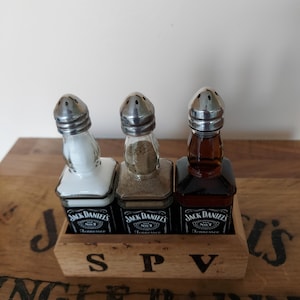 May include: A trio of Jack Daniel's-branded salt and pepper shakers in a wooden holder. The glass shakers have metal tops and contain white, tan, and brown spices. The wooden holder is marked with the letters "S P V".