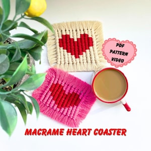 以下が含まれることがあります： クリーム色の赤いハートとピンク色の赤とピンクのハートの2つのマクラメハートコースター。コースターは、コーヒーカップと「PDF PATTERN VIDEO」と書かれた看板のある白い表面にあります。