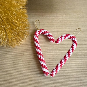 Puede incluir: Un par de pendientes en forma de bastón de caramelo rojo y blanco con ganchos dorados. Los pendientes están hechos de hilo trenzado y tienen forma de corazón.