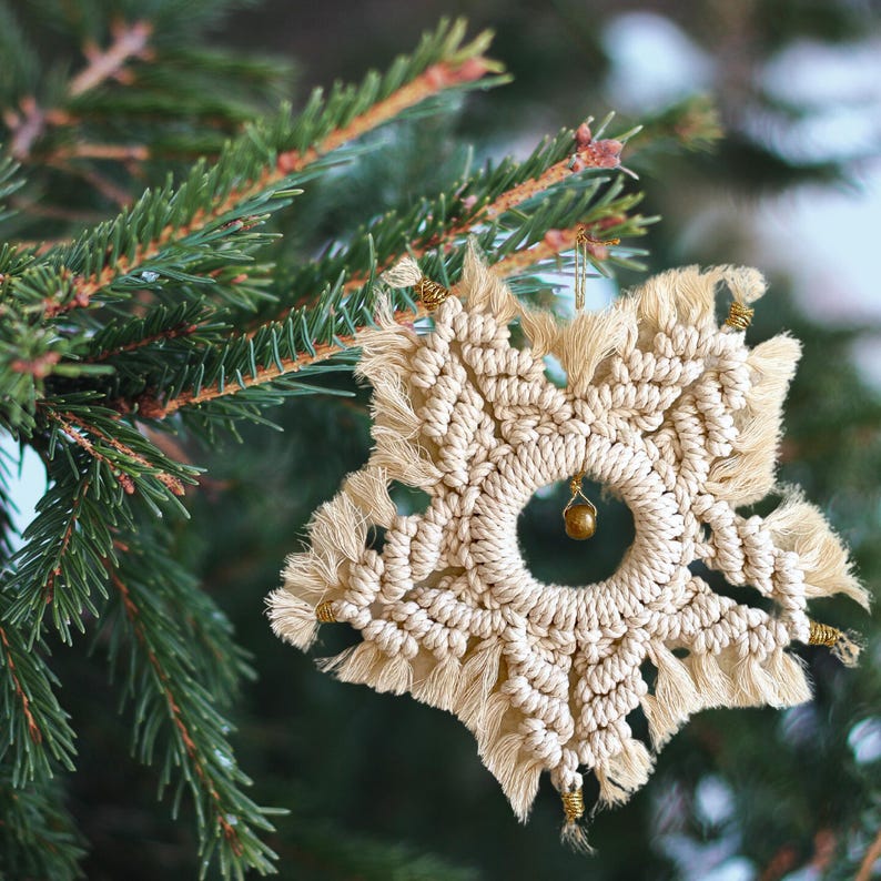 Peut inclure: Une d&eacute;coration de No&euml;l en forme de flocon de neige en macram&eacute; blanc avec une cloche dor&eacute;e suspendue au centre. La d&eacute;coration est suspendue &agrave; une branche de pin verte.