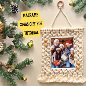 May include: A macrame wall hanging with a photo frame and wooden beads. The frame contains a photo of a group of people smiling and wearing Santa hats. The text "MACRAME XMAS GIFT PDF TUTORIAL" is written in yellow on a white background.
