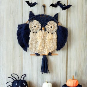 Op de afbeelding: Een marineblauwe en cr&egrave;mekleurige macram&eacute; uil wanddecoratie met een houten zitstok. De uil heeft grote, cirkelvormige ogen en gevederde vleugels. Twee zwarte vleermuisdecoraties bevinden zich boven de uil. Een kleine oranje pompoen en witte pompoen staan eronder.