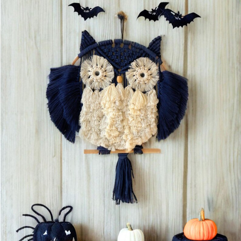 Op de afbeelding: Een marineblauwe en cr&egrave;mekleurige macram&eacute; uil wanddecoratie met een houten zitstok. De uil heeft grote, cirkelvormige ogen en gevederde vleugels. Twee zwarte vleermuisdecoraties bevinden zich boven de uil. Een kleine oranje pompoen en witte pompoen staan eronder.