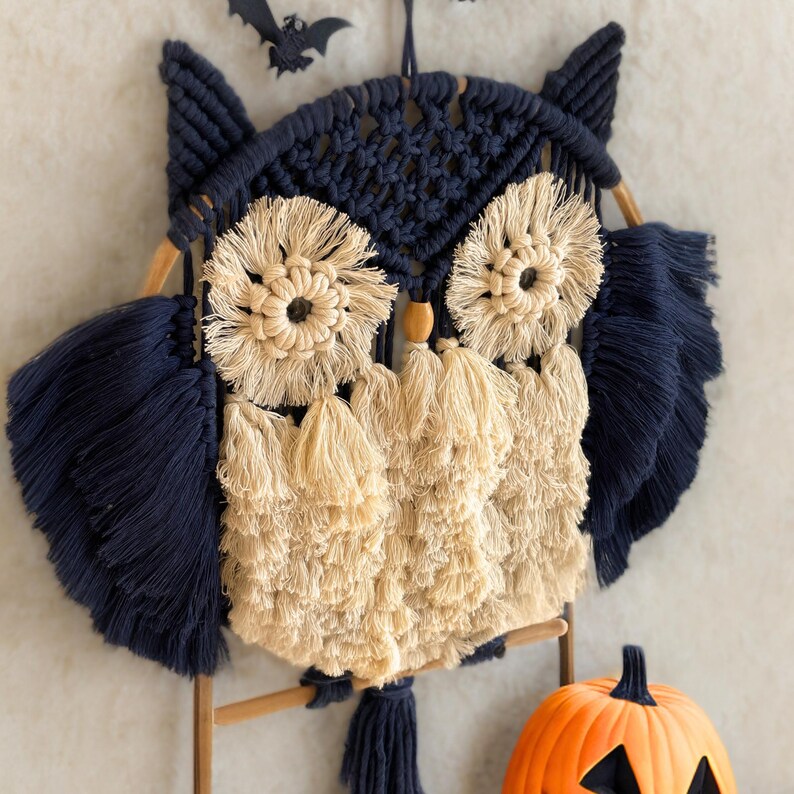 Op de afbeelding: Een macram&eacute; uil wanddecoratie in marineblauw en beige. De uil heeft grote, cirkelvormige ogen en veervormige details. Een houten deuvel en een kleine oranje pompoen met een uitgesneden gezicht zijn ook zichtbaar.
