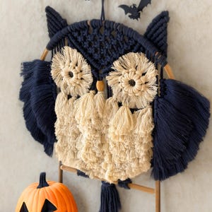 Op de afbeelding: Een macram&eacute; uil wanddecoratie in marineblauw en beige. De uil heeft grote, cirkelvormige ogen, gevederde vleugels en een kwastachtig lichaam. Een kleine, oranje pompoen en een zwarte vleermuisdecoratie zijn ook zichtbaar.