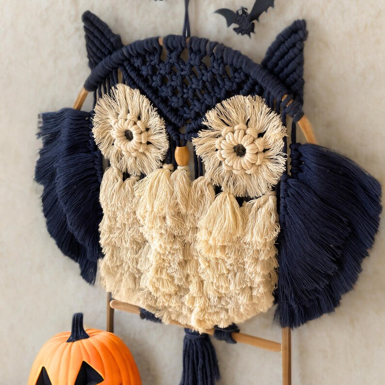 Op de afbeelding: Een macram&eacute; uil wanddecoratie in marineblauw en beige. De uil heeft grote, cirkelvormige ogen, gevederde vleugels en een kwastachtig lichaam. Een kleine, oranje pompoen en een zwarte vleermuisdecoratie zijn ook zichtbaar.