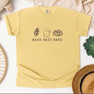 May include: Yellow t-shirt with the words "MAKE WAIT BAKE" and illustrations of wheat, a jar, and a loaf of bread. The shirt is made of a soft material and has short sleeves.